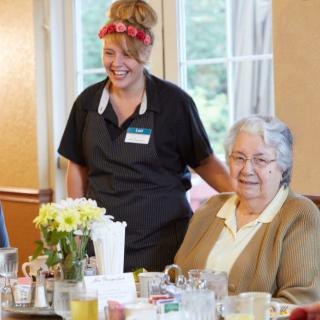 A caregiver and an elderly person smiling together at a dining table in a nursing home.