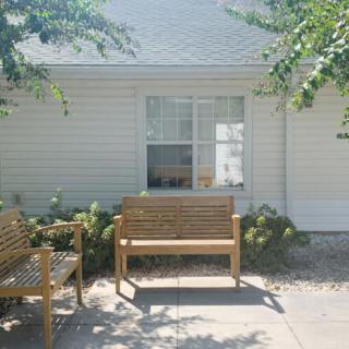 A sunny outdoor seating area in front of a house with wooden benches and trees providing shade.