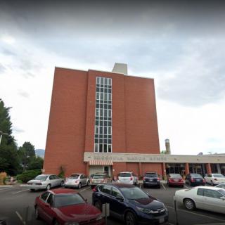 A multi-story brick building with a parking lot filled with cars under a cloudy sky.