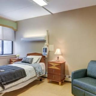 A hospital room featuring a bed, bedside table with a lamp, and a chair.