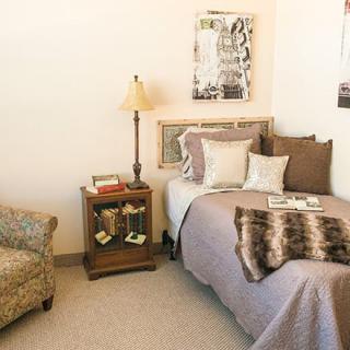 A cozy bedroom featuring a single bed with pillows and a blanket, an elegant nightstand with a lamp, and an armchair.