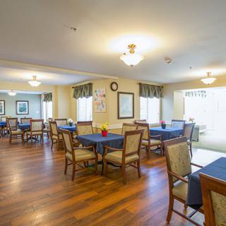 A spacious dining room with neatly arranged tables and chairs on a wooden floor, illuminated by ceiling lights and natural light from large windows.