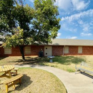 A brick house with a yard featuring a tree and picnic bench under a blue sky.