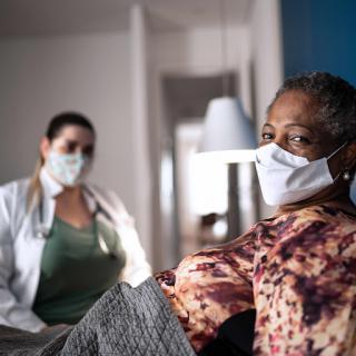 A healthcare professional in a mask attending to an elderly patient in a cozy indoor setting.