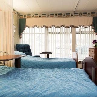 A hospital room with two blue-covered beds and large windows with blinds.