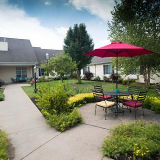 A scenic outdoor garden area with a patio, featuring a red umbrella and seating surrounded by lush greenery.