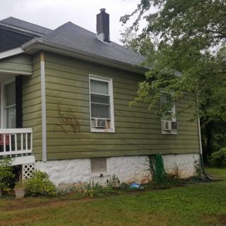 A small green-sided house with a porch and grass yard, surrounded by trees.