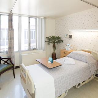 A well-lit hospital patient room featuring a bed, a small table, and a chair beside the window.