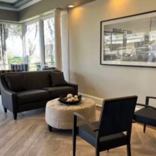 A modern living room with a sofa, chairs, and a central coffee table, featuring large windows and wall art.