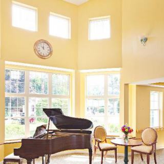A bright living room with a grand piano, yellow walls, and abundant sunlight streaming through large windows.