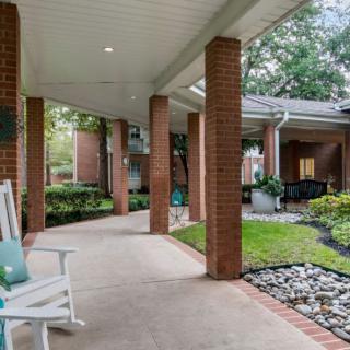 A peaceful outdoor patio with a covered walkway, surrounded by greenery and featuring brick columns and a rocking chair.