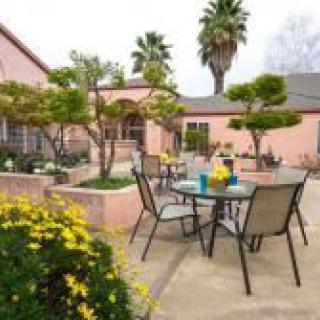 An inviting outdoor patio area with tables and chairs surrounded by lush greenery and a pink house in the background.