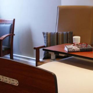 A cozy hospital room with chairs, a bed, and a table with books and a cup.