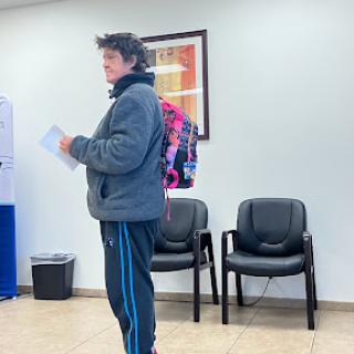 A person stands in a waiting room, holding documents and wearing a backpack.