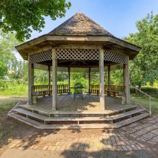 A wooden gazebo surrounded by greenery on a sunny day.