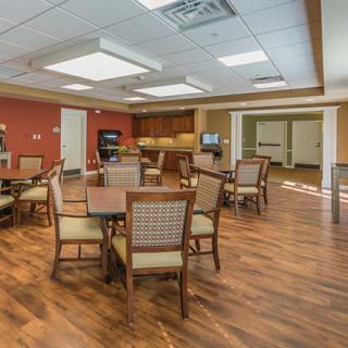 A spacious dining area with multiple tables and chairs, featuring a wooden floor and a kitchenette in the background.