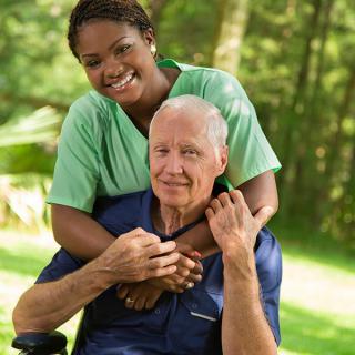 A caregiver embracing an elderly person in a wheelchair outdoors.