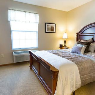 A cozy bedroom with a neatly made bed, bedside lamp, and natural light coming through the window.