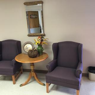 A cozy seating area featuring two purple chairs, a round wooden table with decorative items, a small vase of flowers, and a mirror on the wall.