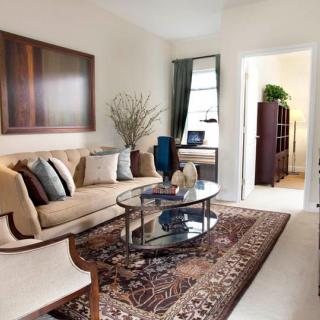 A cozy living room with a beige sofa, glass coffee table, and an intricate rug, complemented by wall artwork and window curtains.