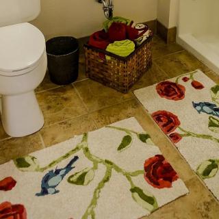 A bathroom with a floral and bird patterned bath mat, tiled floor, and a basket containing towels.
