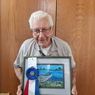 An elderly man proudly holds a framed painting adorned with a blue ribbon award.