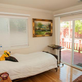 A cozy bedroom with a bed, painting on the wall, and sunlight streaming through a sliding glass door.