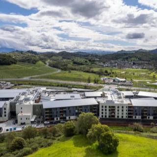 Aerial view of modern buildings surrounded by lush greenery and hills under a cloudy sky.