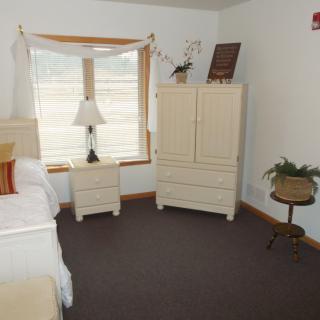 A cozy bedroom featuring a bed, nightstand with a lamp, and a dresser near the window.