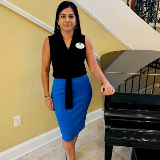 A woman in a stylish black top and blue skirt poses elegantly beside a piano.