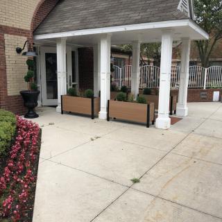 A building entrance with white columns and planters, surrounded by a flower-lined sidewalk.