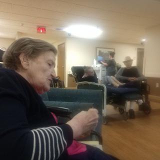 An elderly woman sitting in a nursing home, with others and caregivers in the background.
