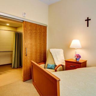 A cozy bedroom featuring a bed, chair, and a lamp, with a cross on the wall.