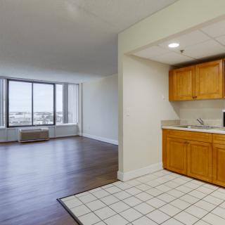 An interior view of an apartment featuring a kitchen with wooden cabinets and a spacious living area with large windows and hardwood flooring.