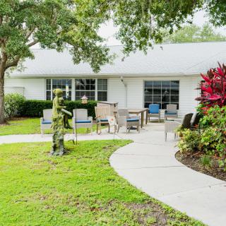 A serene garden setting with a curved walkway leading to outdoor seating in front of a house, surrounded by trees and well-maintained landscaping.