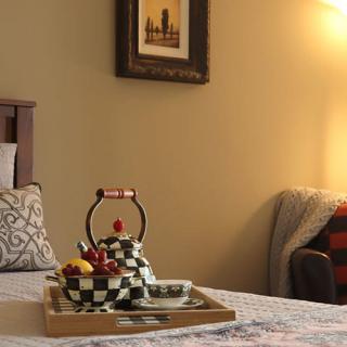 A cozy bedroom scene featuring a decorative teapot on a tray, with soft lighting from a lamp and artwork on the wall.