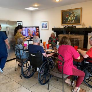 A group of seniors in wheelchairs gathered around a table in a nursing home, with nurses assisting and a television playing.