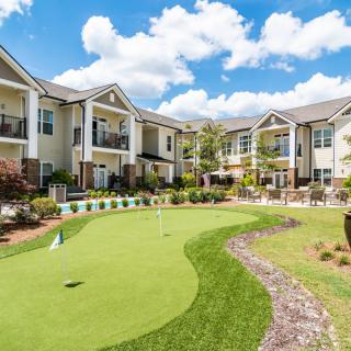 A modern apartment complex with a well-maintained golf putting green in the courtyard on a sunny day.