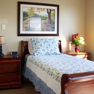 A cozy bedroom with a single bed featuring floral bedding, a nightstand with a lamp, and a framed painting above the bed.