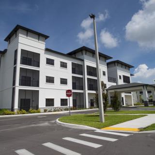 A modern, multi-story building with a landscaped exterior and adjacent road under a cloudy sky.