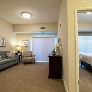 A cozy apartment interior featuring a living room with a sofa and lamp, leading into an adjoining bedroom.