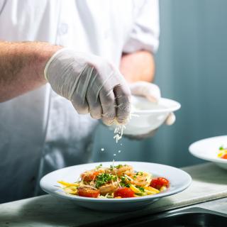 A chef garnishing a pasta dish with herbs in a restaurant kitchen.