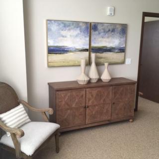 A cozy room corner with a wooden cabinet, decorative vases, and landscape artworks above, accompanied by a cushioned chair.