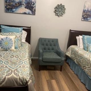A cozy bedroom with two twin beds and a teal armchair, featuring patterned bedding and wall art.