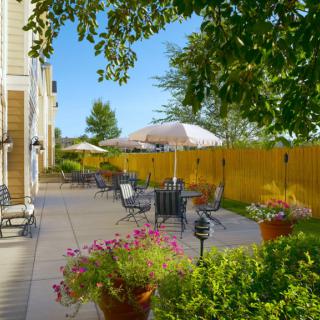 A sunny patio with outdoor seating, tables, and umbrellas surrounded by a garden and a wooden fence.