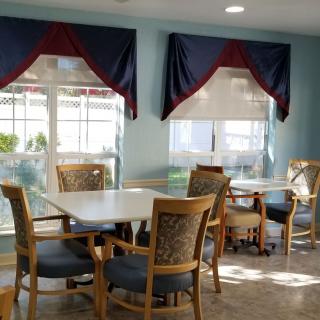 A sunlit dining area with tables and chairs, adorned with curtains over large windows.