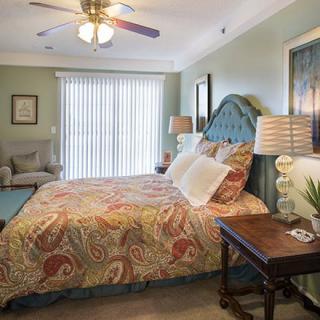 A cozy and well-decorated bedroom featuring a large bed with colorful bedding, a nightstand with a lamp, and a comfortable chair. A ceiling fan hangs overhead, and sunlight filters through vertical blinds.