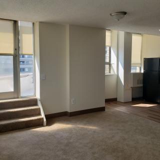 An empty apartment interior with carpet flooring, natural light streaming through large windows, and a small set of stairs leading to a balcony.