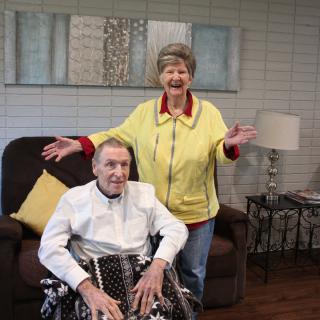 An elderly couple in a living room, with one person sitting in a wheelchair and the other standing beside them, both smiling.