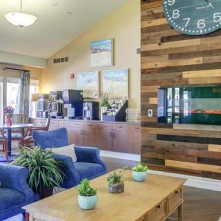 A cozy lounge area with blue chairs, a wooden coffee table, and a coffee station in the background. A large clock is mounted on the wood-paneled wall.